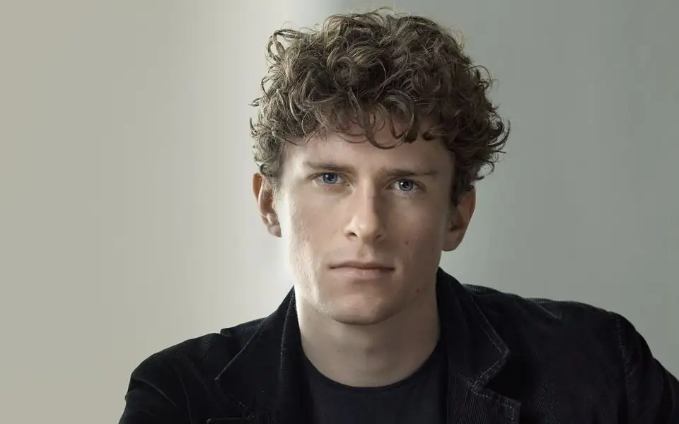 Actor seated on a stool in a portrait studio, softly lit against a grey backdrop, looking into the camera