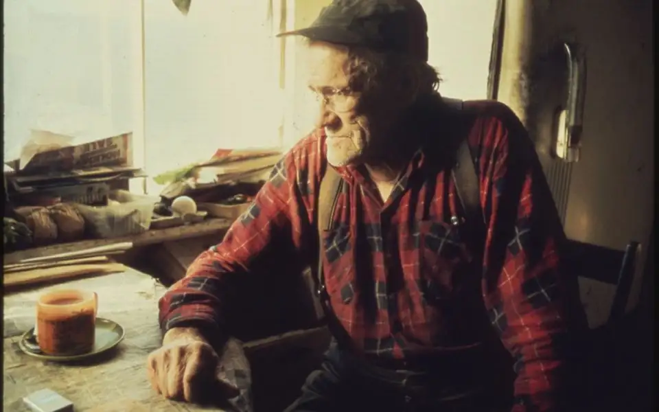 Woodworker standing in her workshop among tools and sawdust, lit by a single window, looking calmly at the camera