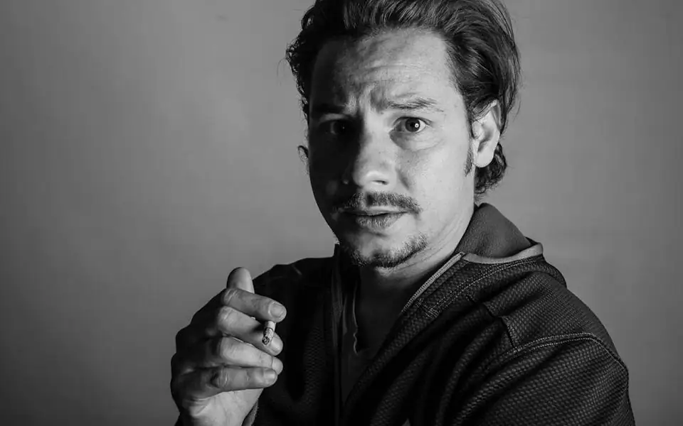 Actor sitting on a stool in front of a grey studio backdrop while a photographer checks a frame on the camera screen